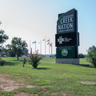 Very large outdoor sign with text 'Muscogee Creek Nation Tribal Complex' in front of a government building with several flags including US, Oklahoma, and Muscogee